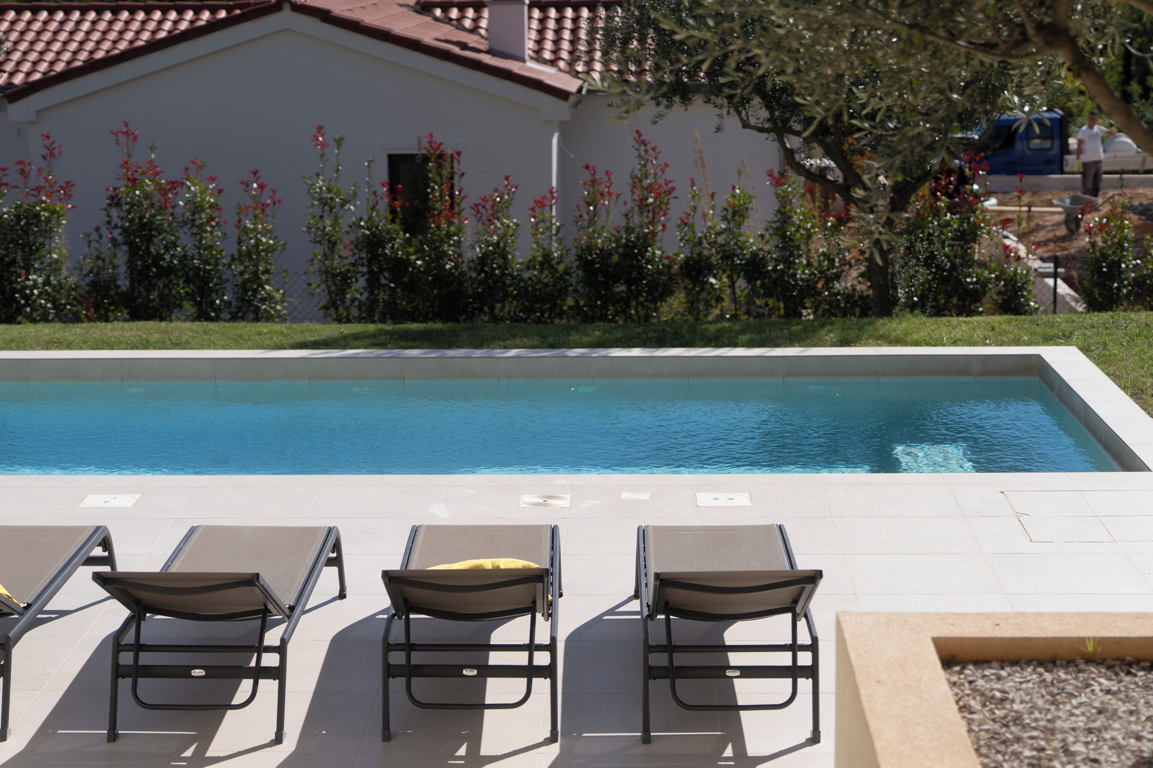 Une piscine avec des chaises longues et un bâtiment surmonté d’un toit recouvert de tuiles rouges.
