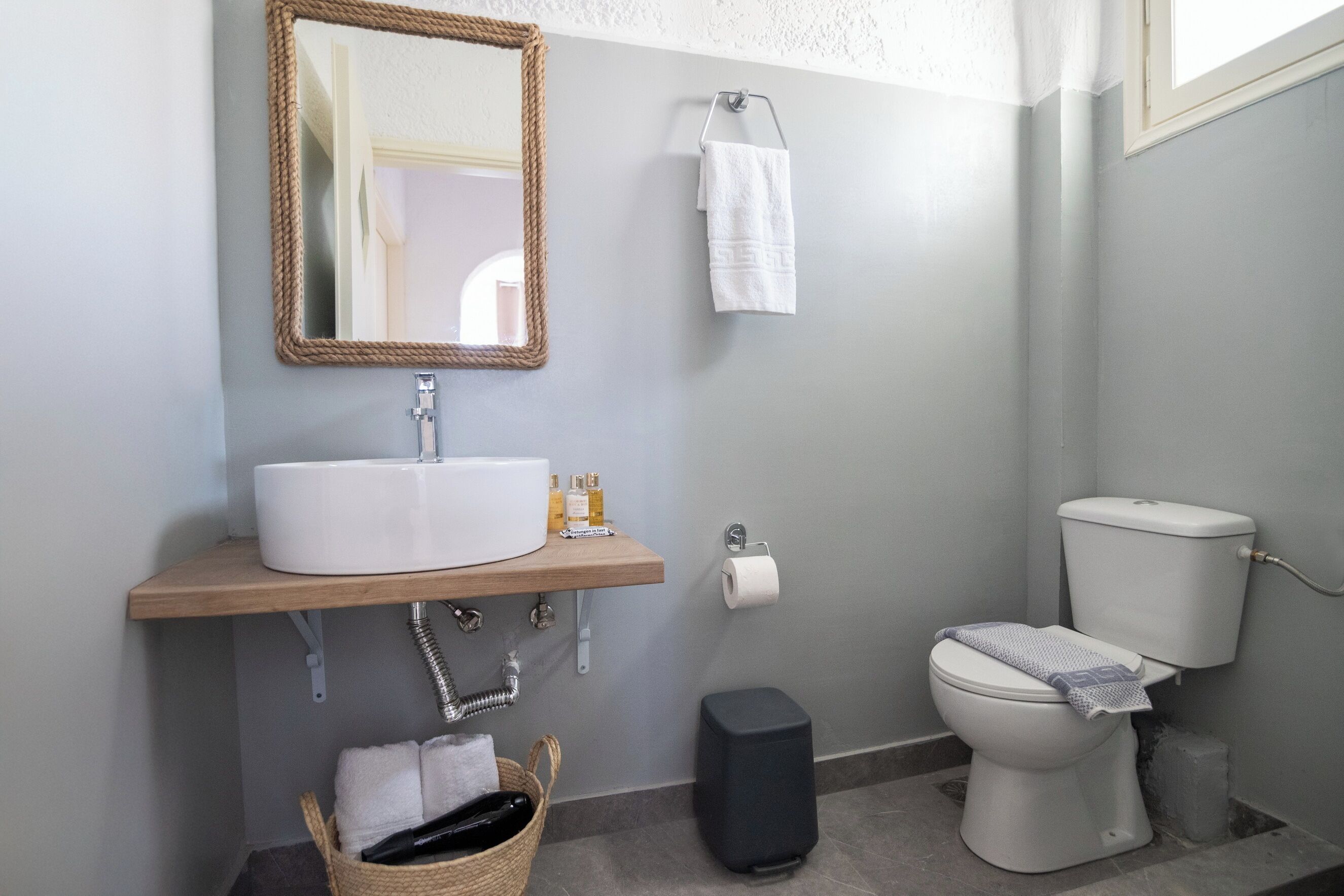 A bathroom with a white sink, mirror, and toilet. There is a towel rack with a towel hanging, a basket with towels, and a trash bin.