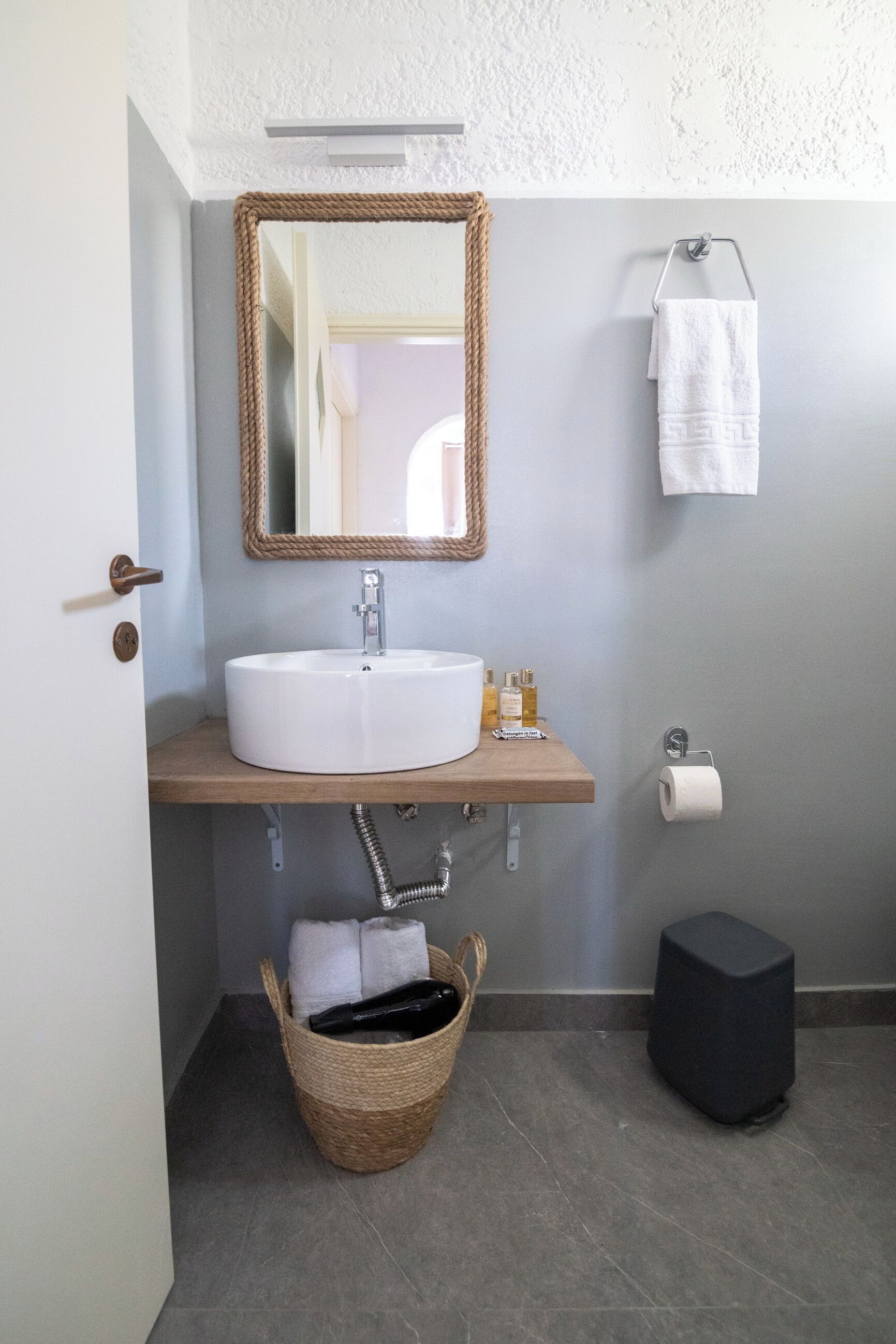 A bathroom with a round sink, a mirror, a towel rack, a toilet paper holder, a trash bin, and a basket.