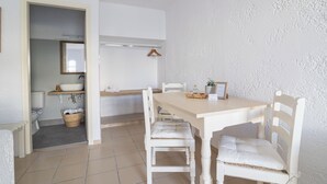 Una habitación pequeña y limpia con una mesa blanca, sillas y un lavamanos. Hay un perchero de madera en la pared y una cesta en el suelo.