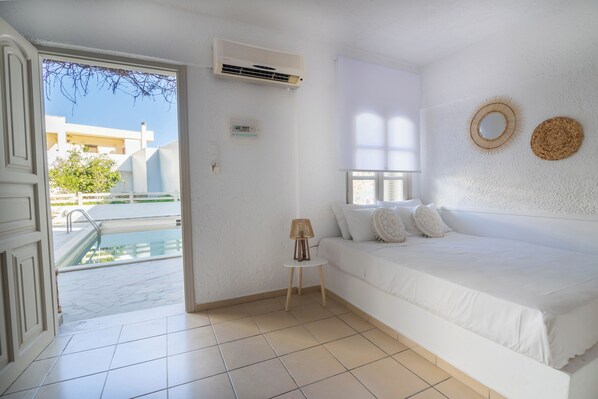Un dormitorio blanco con cama, una mesita de noche con lámpara, una ventana y vistas a una piscina y edificios.
