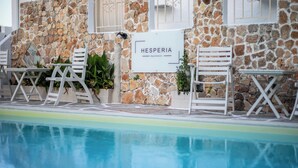 Zona de piscina con paredes de piedra, un cartel que dice "HESPERIA Apartments" y mobiliario exterior blanco.