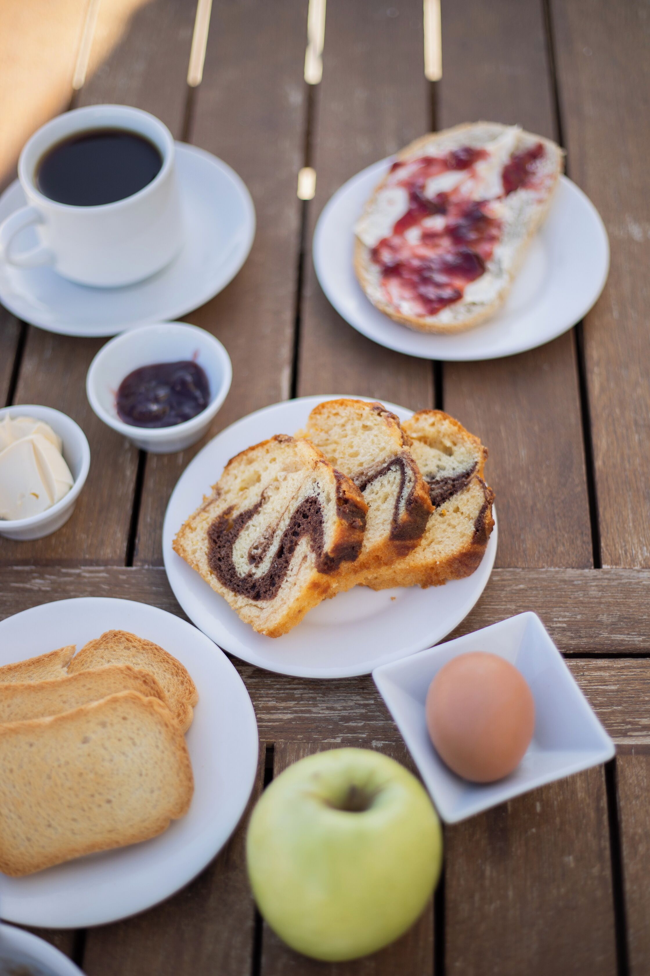 A wooden table with a variety of food items including bread, an apple, a cup of coffee, and a plate with jam.