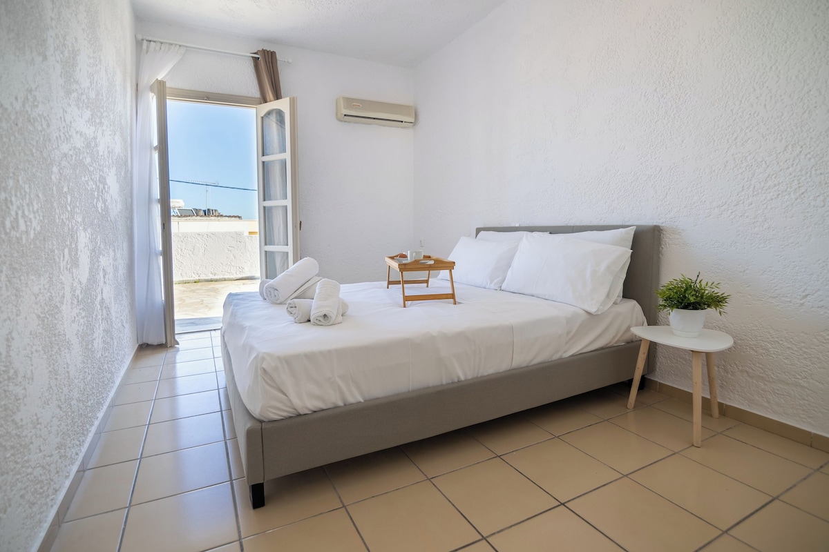 A neatly made bed with white linens, a small bedside table with a plant, and a window with curtains.