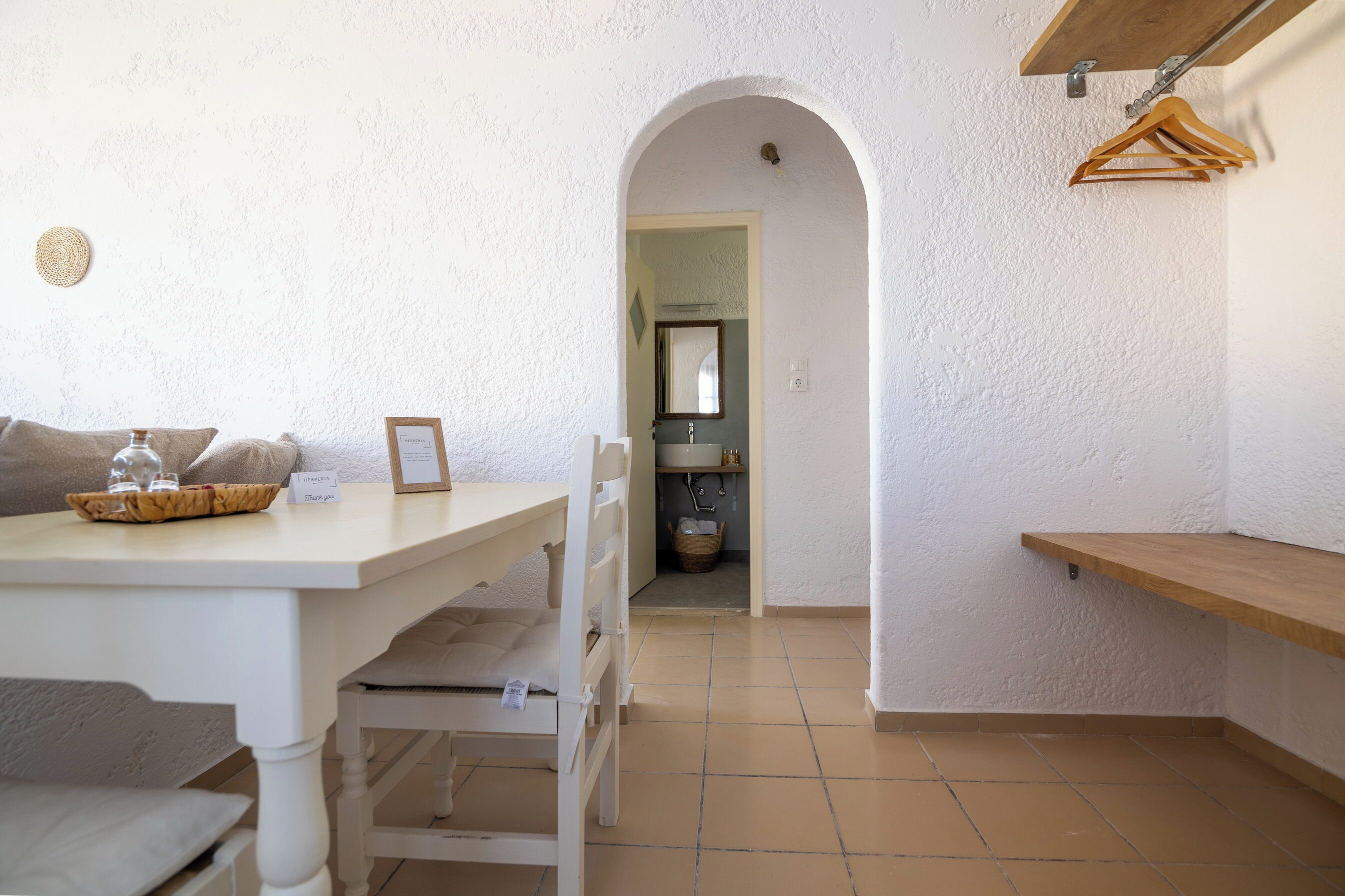 Une pièce de taille modeste, avec une table blanche, un banc et un étagère en bois suspendue.