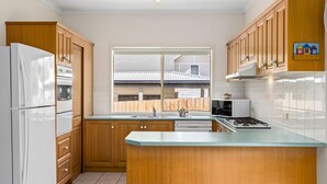 A kitchen with wooden cabinets, a white refrigerator, a stove, and a window overlooking a house. - The Lighthouse Point Lonsdale (Point Lonsdale)