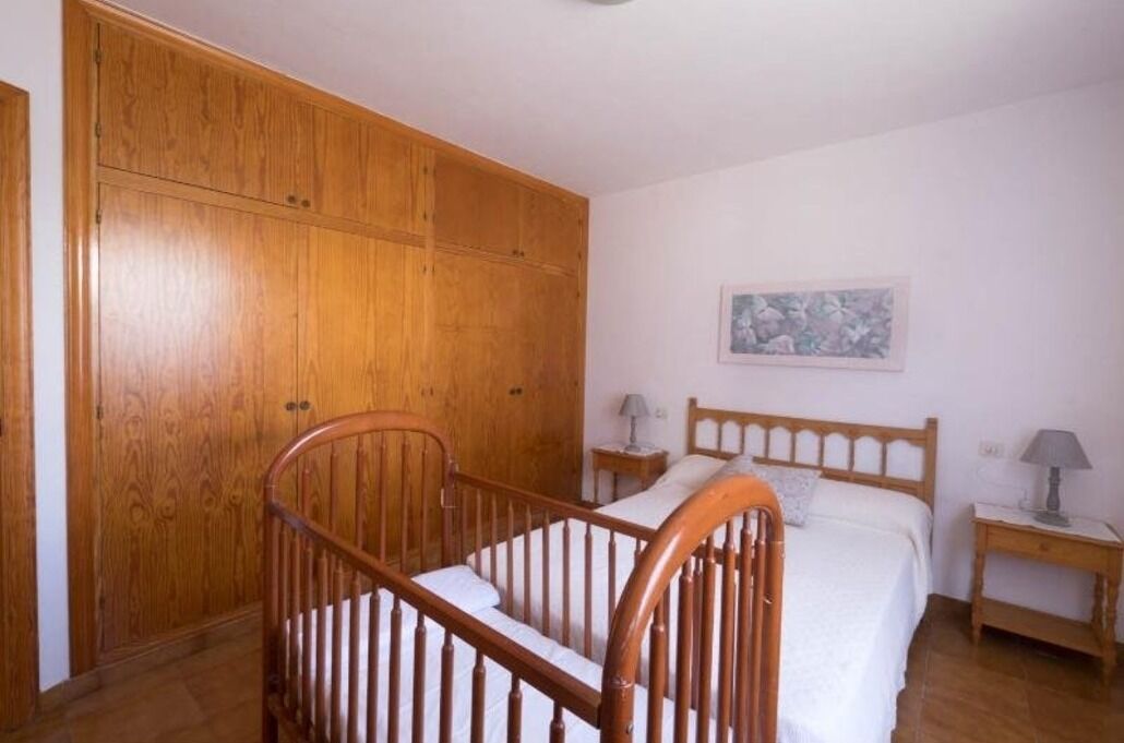 A bedroom with a wooden wardrobe, a single bed, a nightstand, and a wall-mounted lamp.