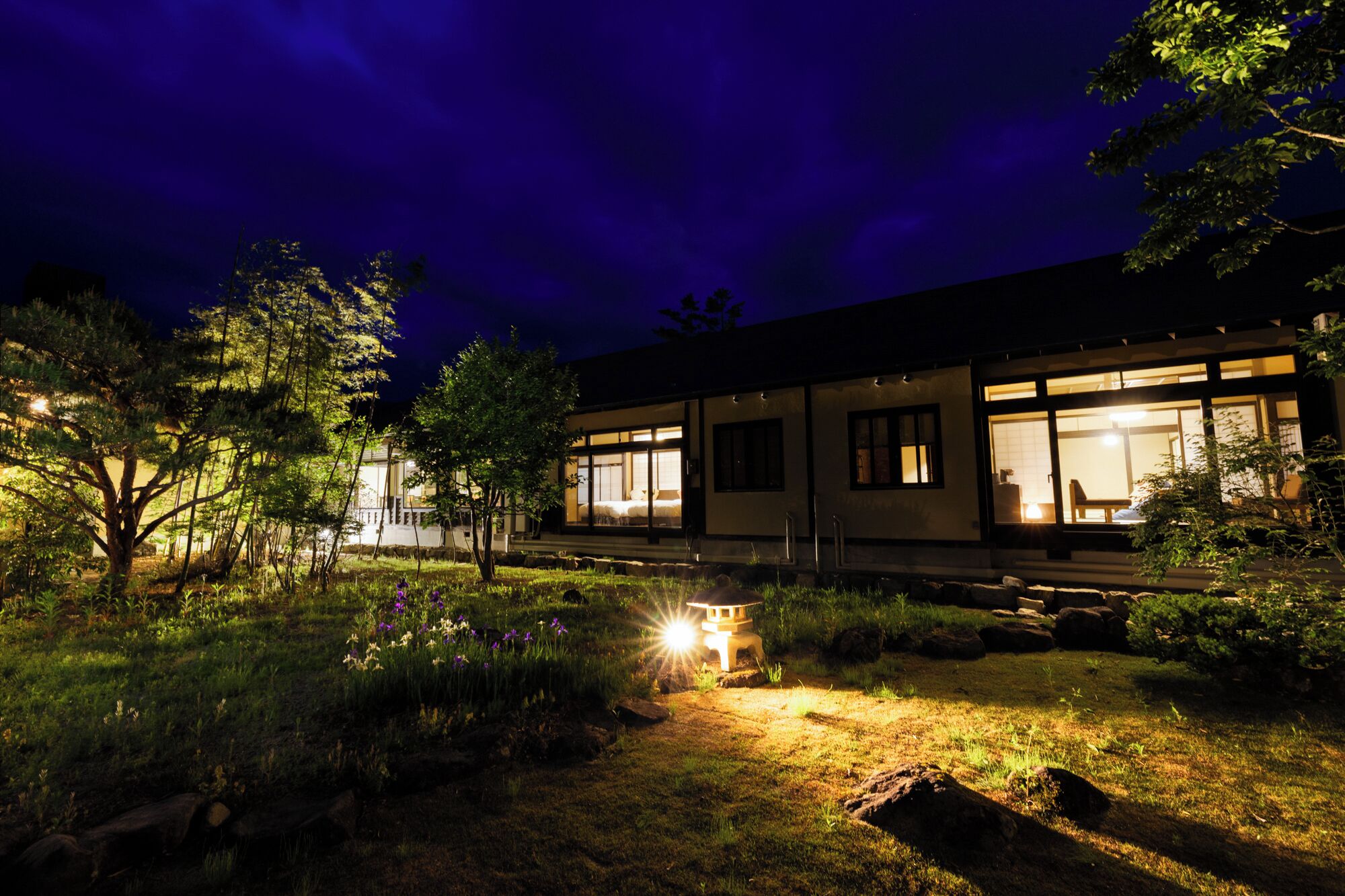 A modern house with large windows and a well-lit garden at night.