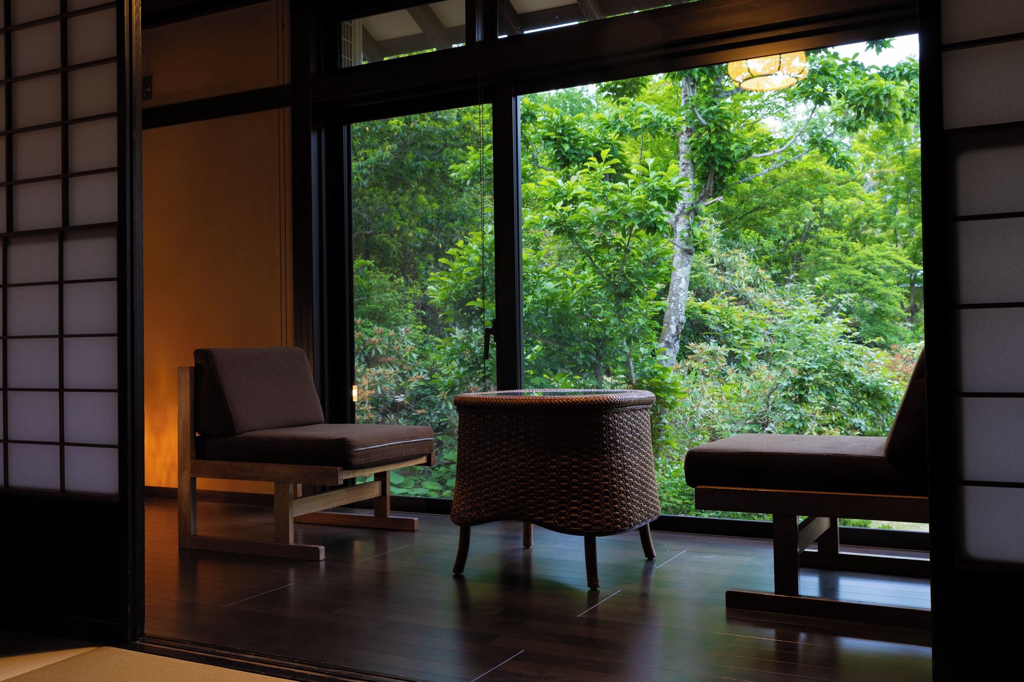 A room with two chairs and a woven stool, large windows with a view of dense greenery.