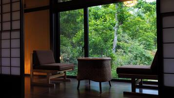A room with two chairs and a woven stool, large windows with a view of dense greenery.
