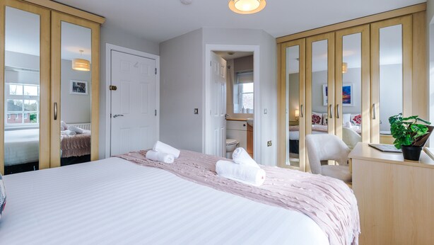 Une chambre d’hôtel avec un grand lit, un bureau, une chaise et un miroir. Il y a une porte qui donne sur une autre pièce et une fenêtre avec vue.