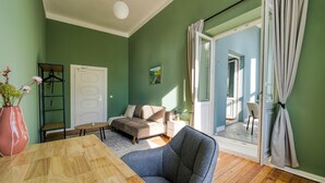 Een kamer met een houten tafel, een blauwe stoel, een bank en een bed. Er is een deur en een raam met gordijnen.