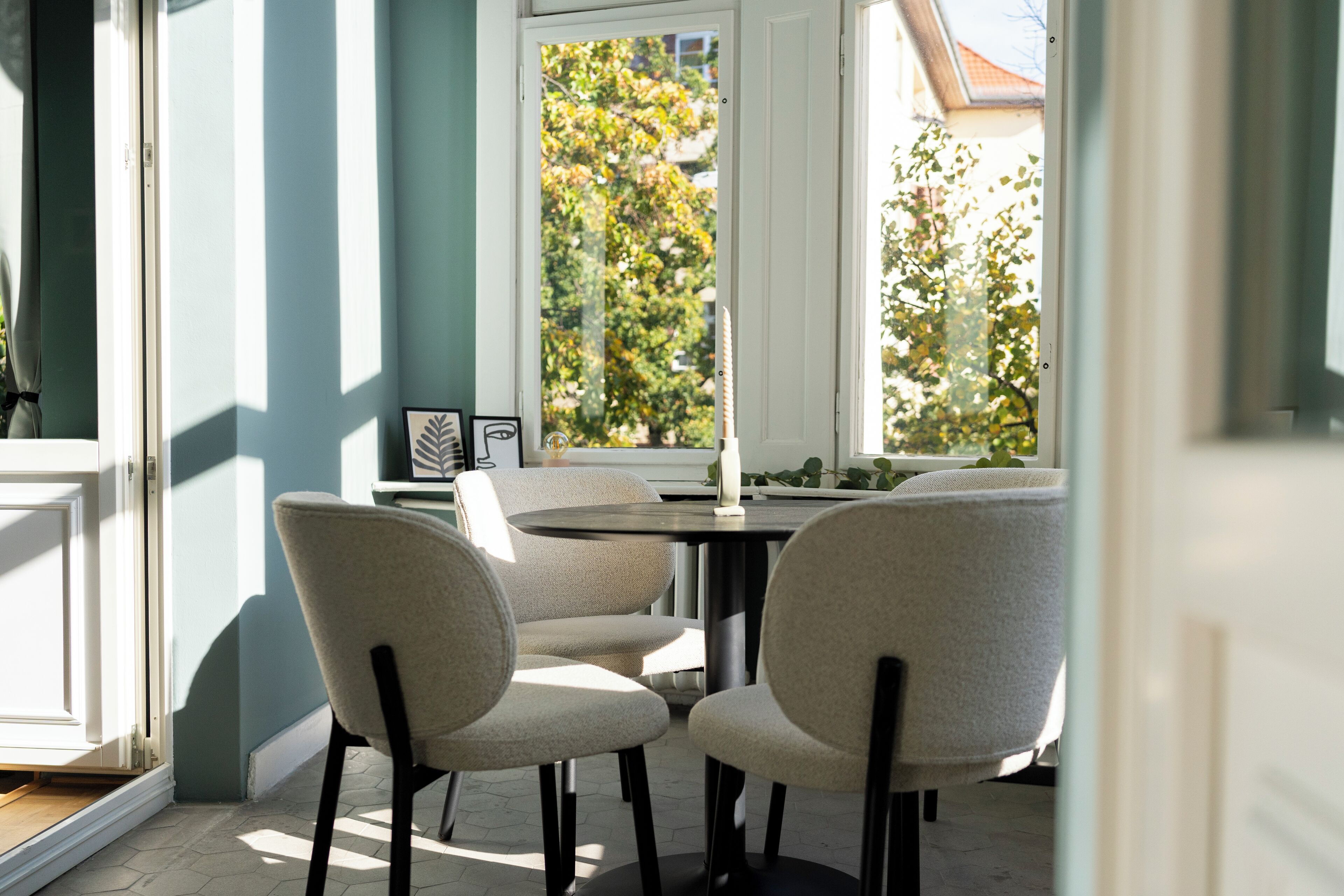 Comedor con una mesa redonda y cuatro sillas, una ventana que da a árboles, y una puerta a la izquierda.