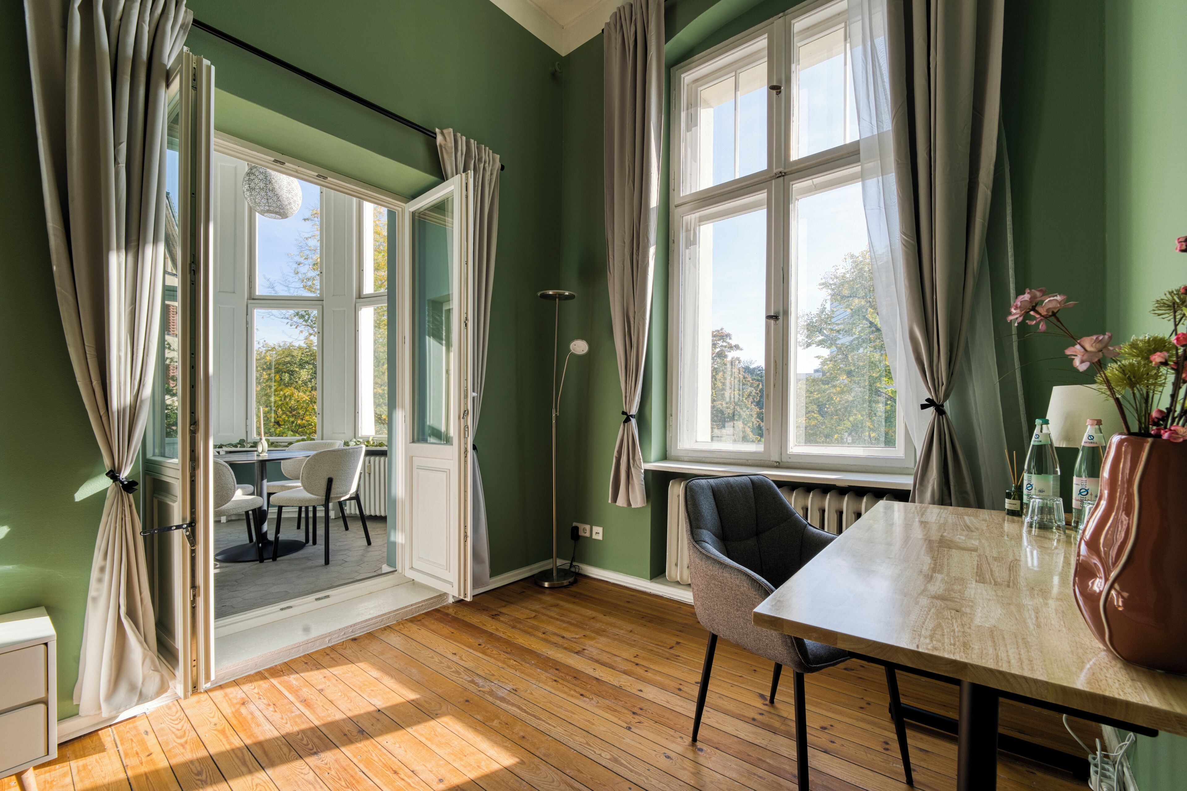 Una habitación con piso de madera, una mesa con sillas, un radiador y una ventana con cortinas.