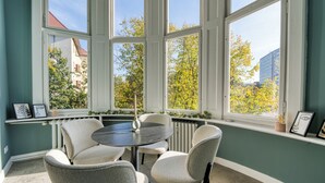 Een kamer met een ronde tafel en vier stoelen, grote ramen met uitzicht op bomen en gebouwen, en een radiator langs de muur.