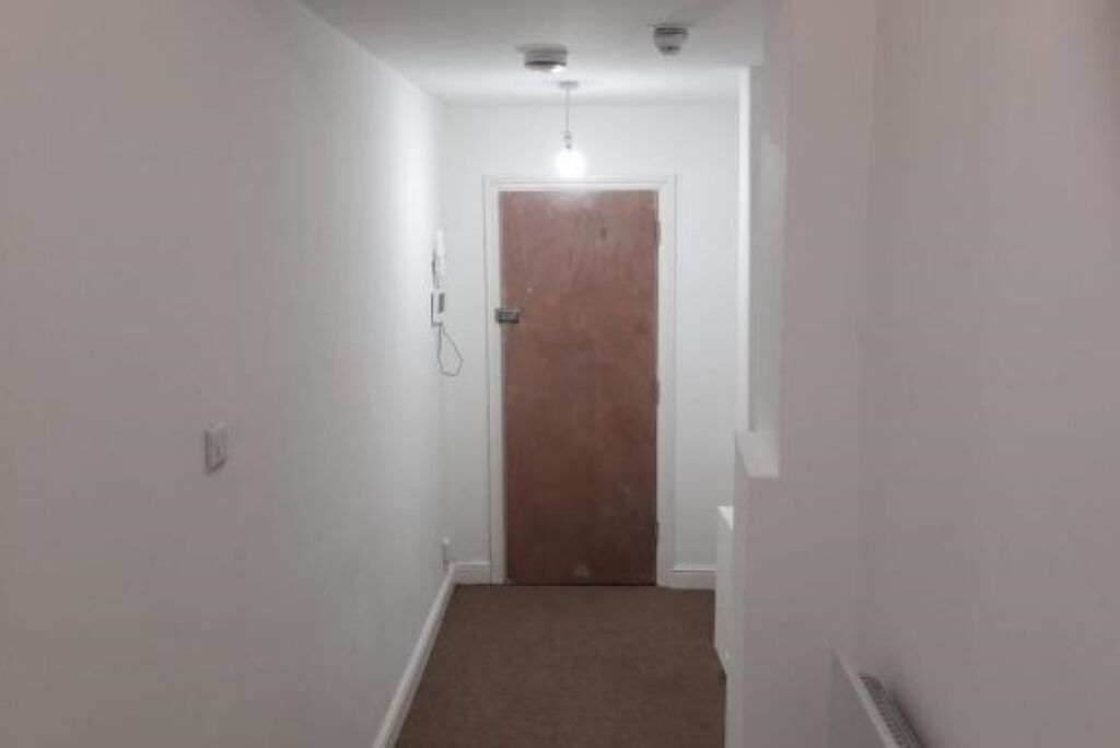 A narrow hallway with a wooden door at the end, white walls, and a light fixture on the ceiling.