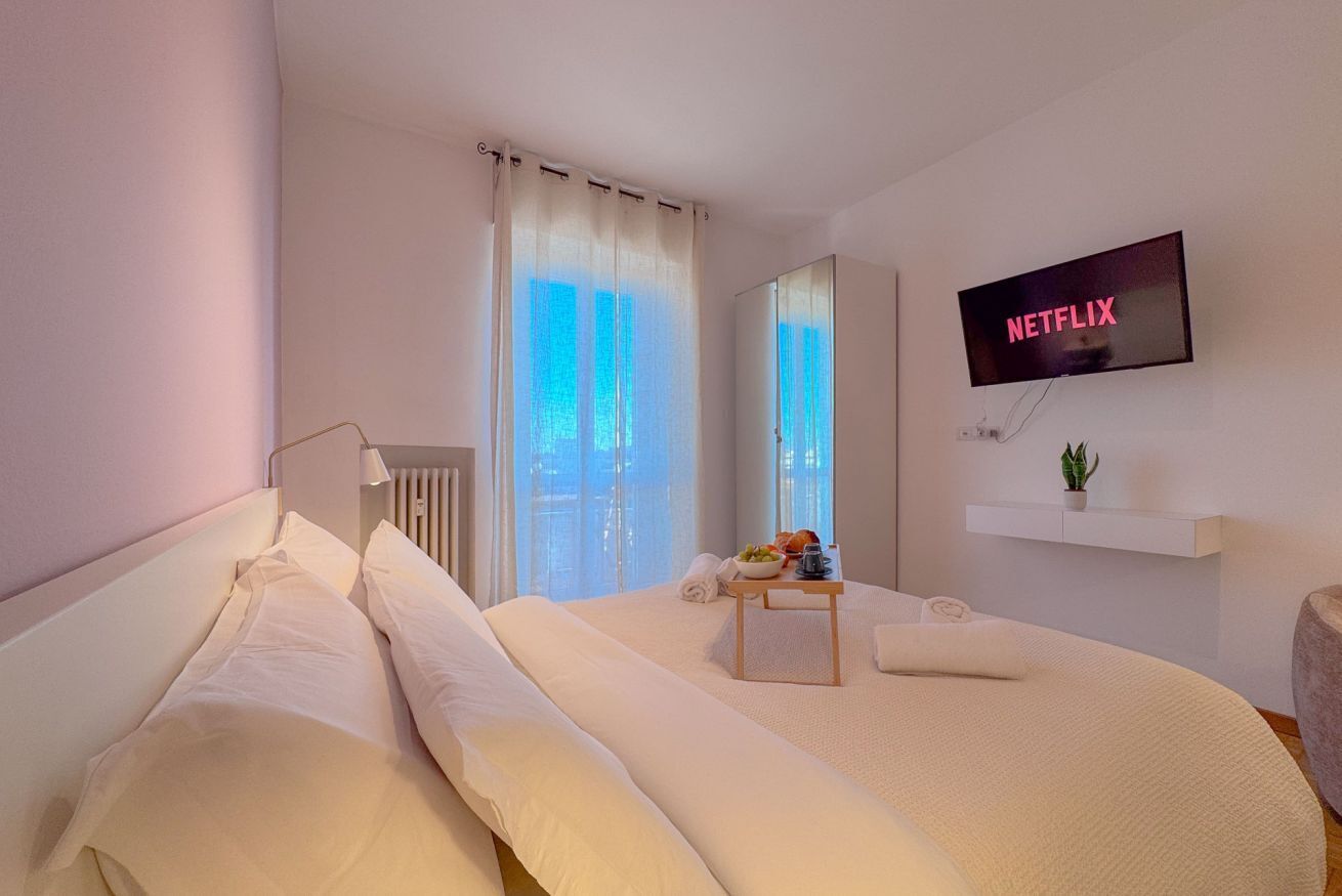Et moderne hotelværelse med en seng, et fjernsyn ophængt på væggen, et lille bord med en skål frugt og en plante på en hylde.