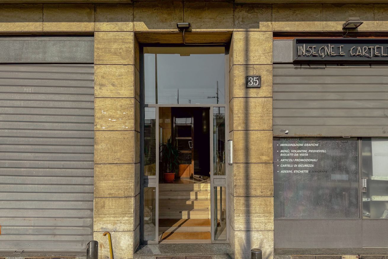 L’entrée d’un immeuble, avec le numéro « 35 » au-dessus de la porte, un volet fermé sur la gauche et une enseigne indiquant des services tels que « IMMAGINAZIONI GRAFICHE » et « ARTICOLI PROMOZIONALI » sur la droite.