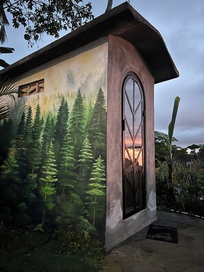 A building with a mural of a forest scene, a large arched window, and a concrete exterior. - Kamanicamp (Mekarsari)