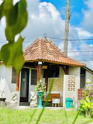 A house with a tiled roof, a porch, and a small garden area with statues and a bench. - Kamanicamp (Mekarsari)