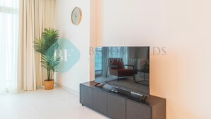 A modern living room with a flat-screen TV on a dark cabinet, a potted plant, a clock on the wall, and a large window with curtains. - Bloomfields Modern 1BR in Thalassa  (Abu Dhabi)
