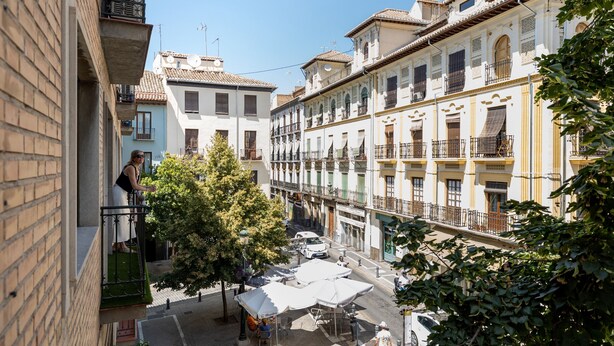 A narrow street with buildings on either side, a tree in the middle, and a person on a balcony.
