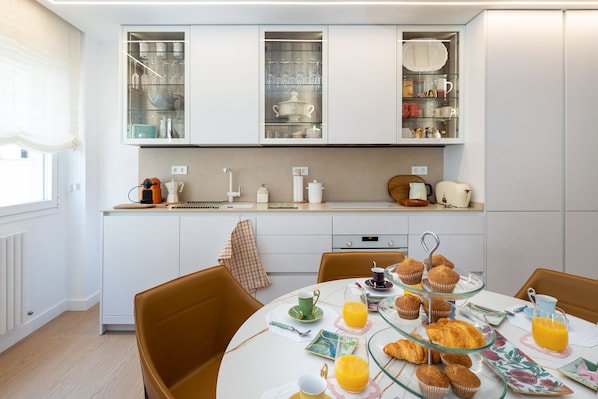 A modern kitchen with white cabinets, a dining area with a round table, and breakfast items like muffins and orange juice.