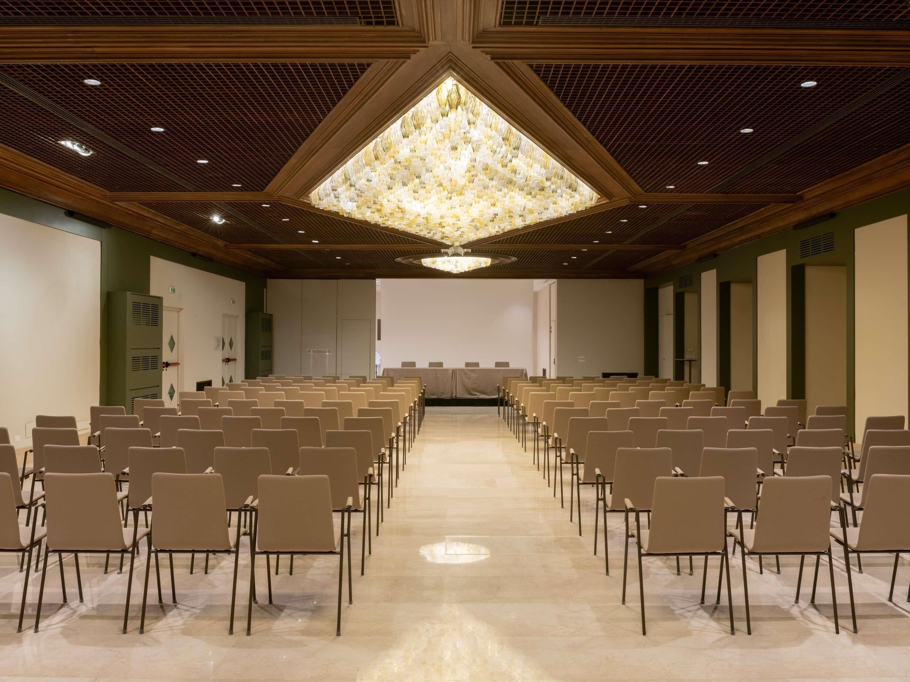 Ein Konferenzraum mit Reihen von Stühlen, einem großen Kronleuchter und einer Projektionsleinwand.