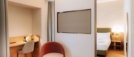 Ett hotellrum med en säng, ett skrivbord med en stol, ett litet runt bord, en röd fåtölj och en stor skärm på väggen.