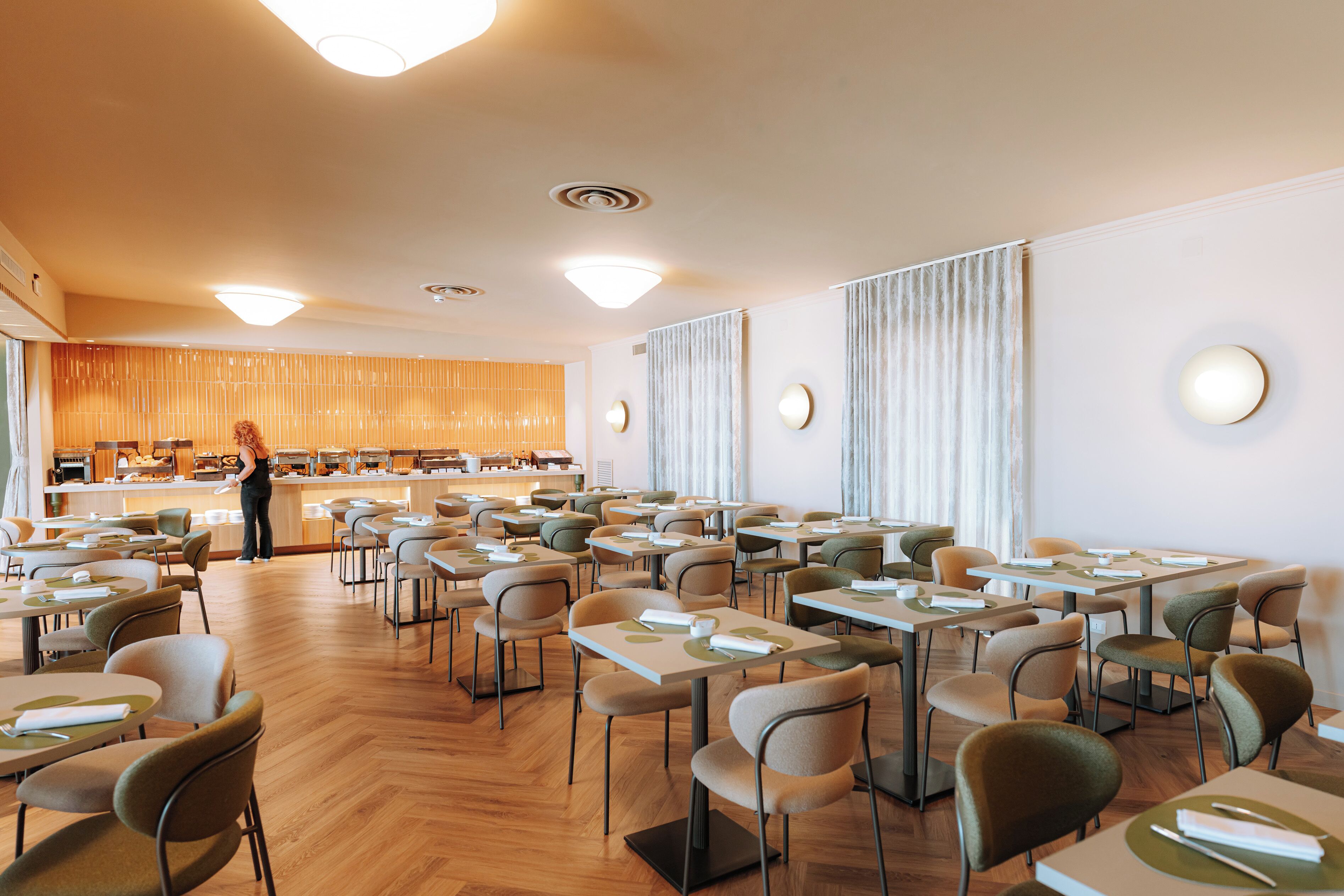 En rummelig spiseplads med flere borde og stole, en disk med en person stående, og et moderne loftdesign.