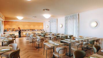 A spacious dining area with multiple tables and chairs, a counter with a person standing, and a modern ceiling design.