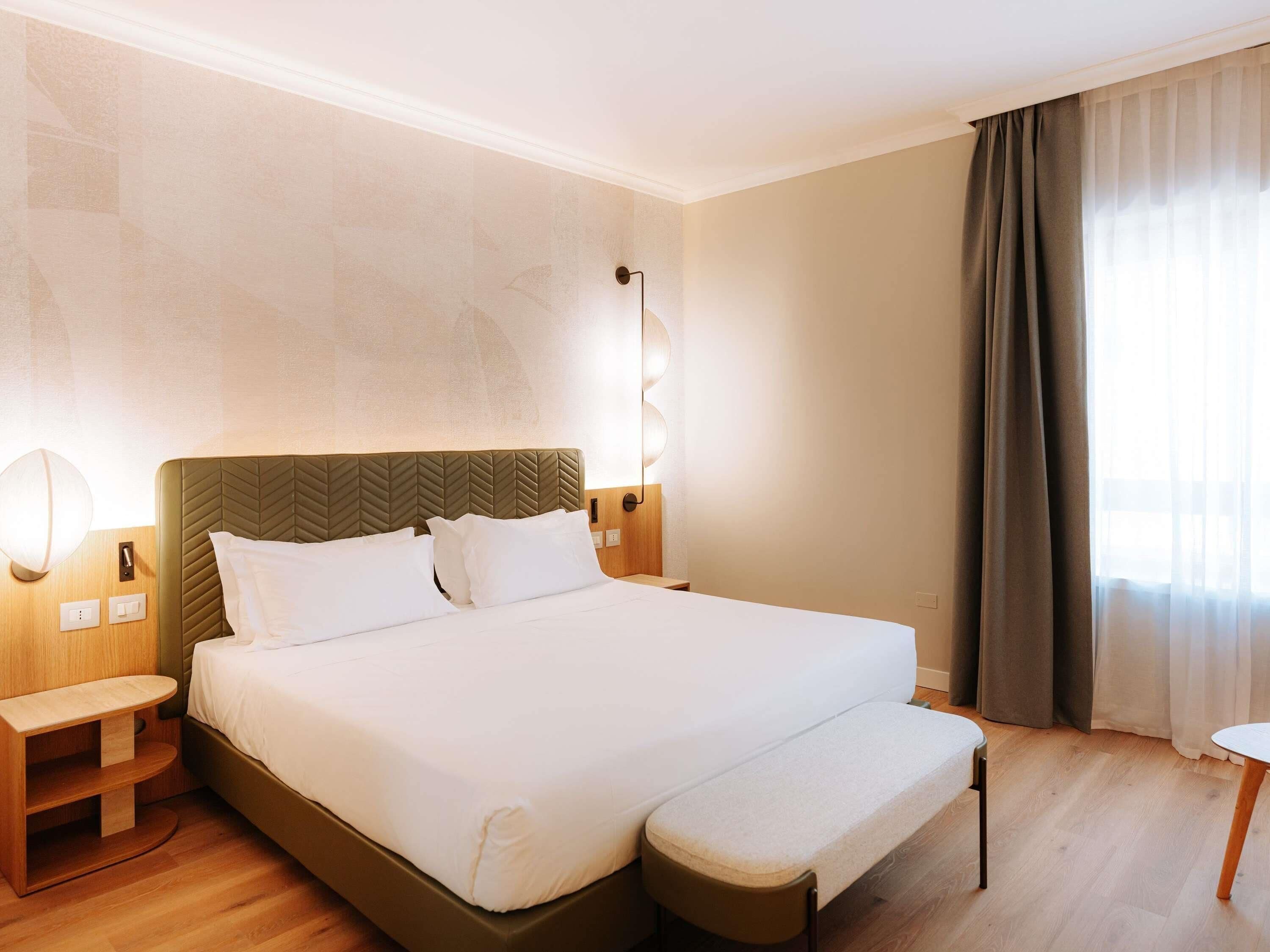 A hotel room with a large bed, bedside tables, a bench, and a window with curtains.