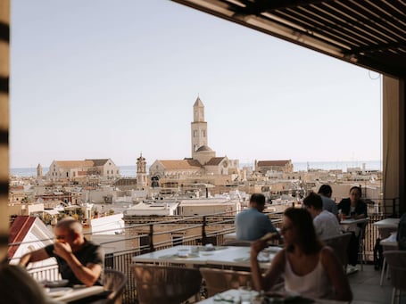 A terrace with tables and chairs overlooking a cityscape with historic buildings and a prominent tower.. Mövenpick Hotel Bari