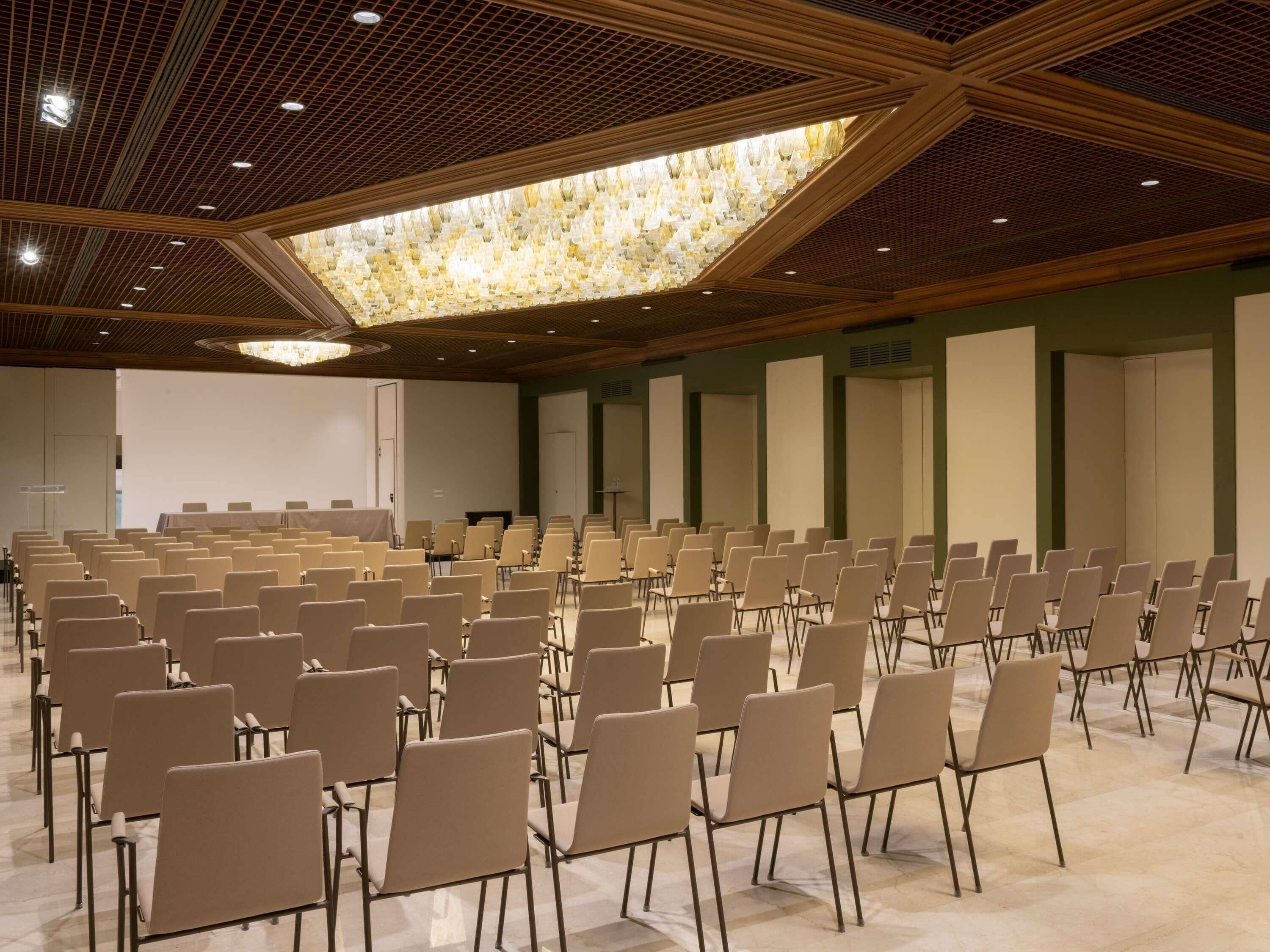Un salón de conferencias con filas de sillas, una gran lámpara de araña y una pantalla de proyector.