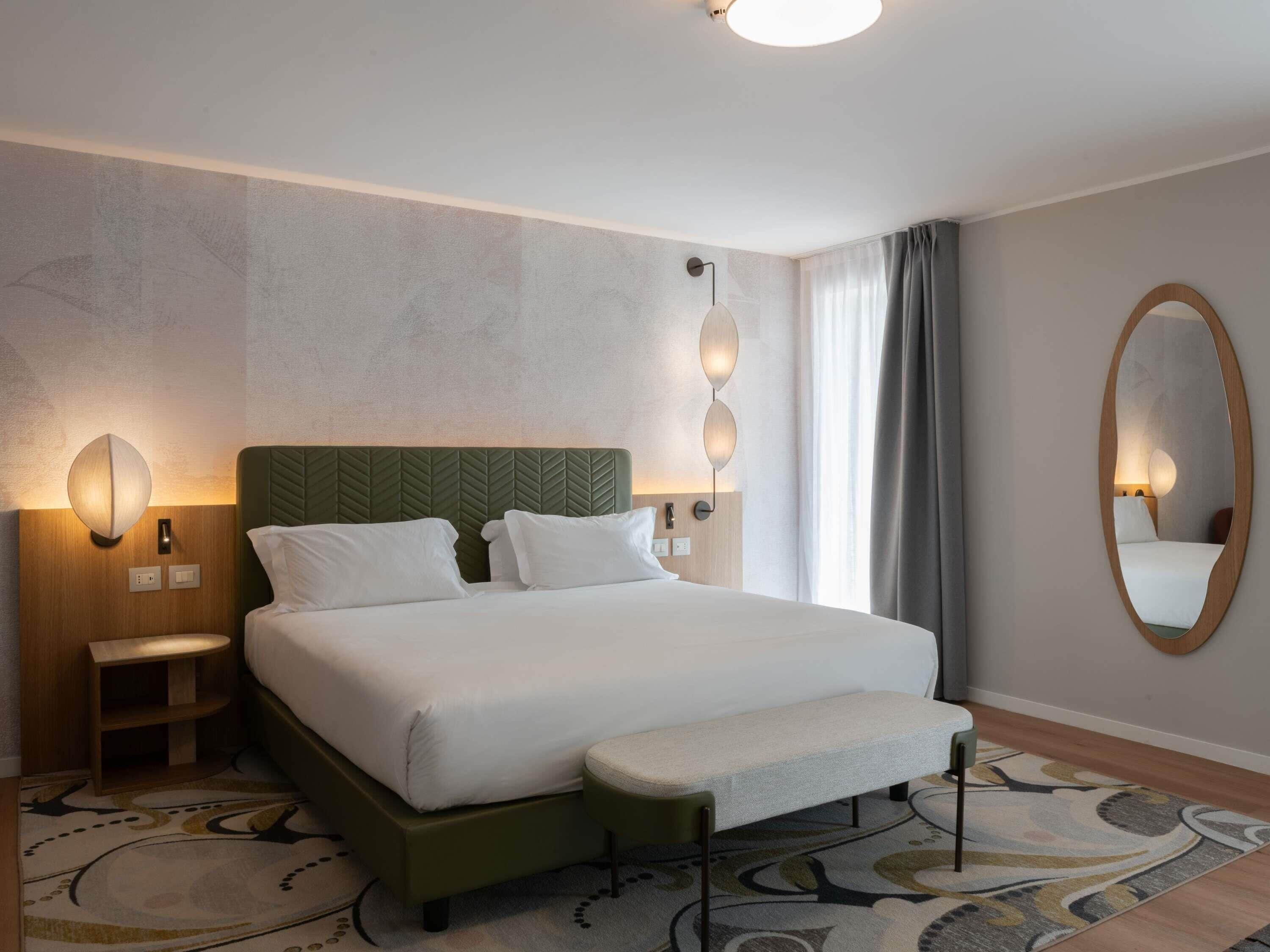 Une chambre d’hôtel avec un lit, une table de chevet, des lampes fixées au mur, un miroir et un tapis à motifs.
