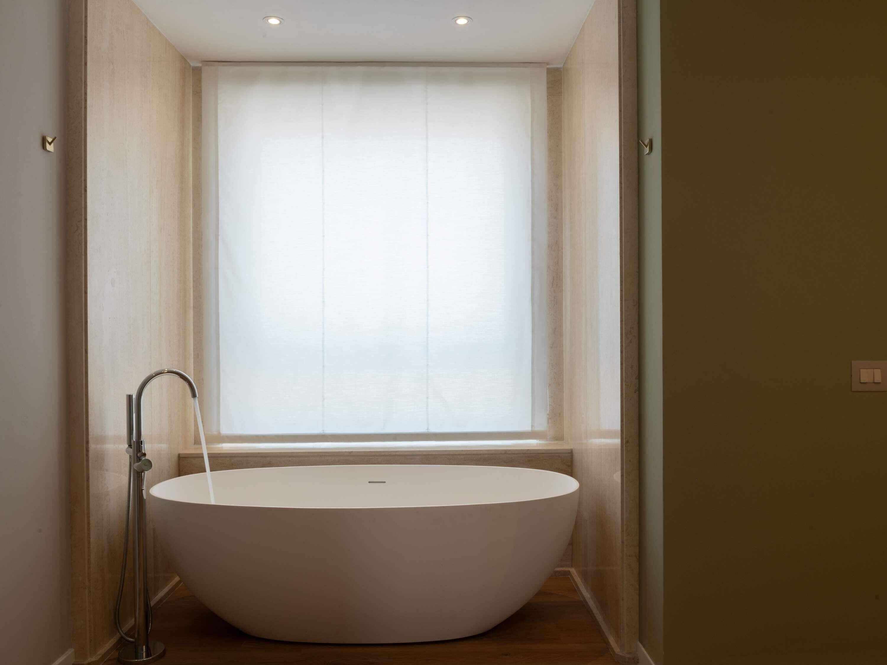 A modern bathroom with a freestanding bathtub, a showerhead, and a window with sheer curtains.