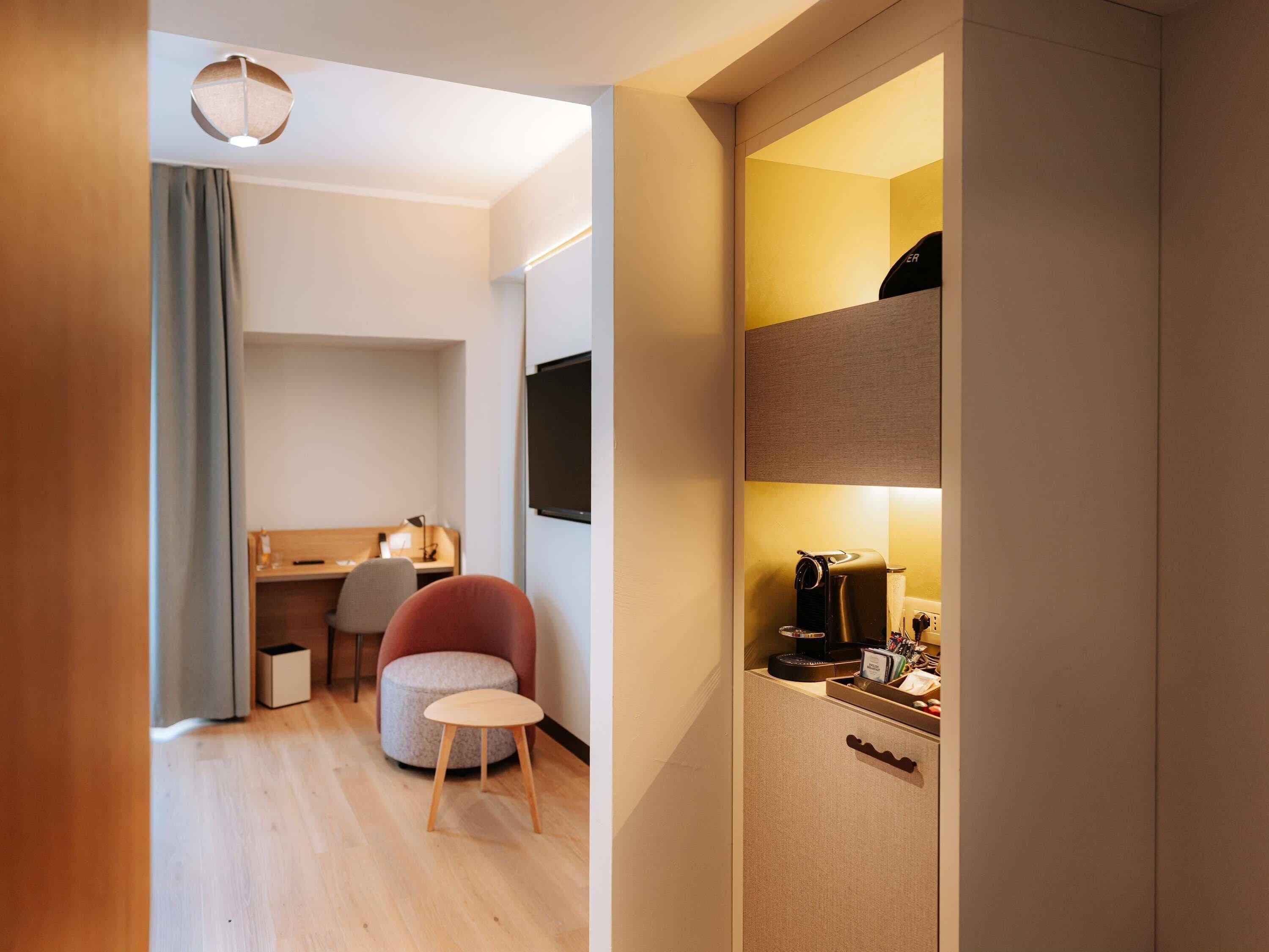 A modern hotel room with a wooden floor, a desk with a chair, a small round table, a wardrobe, and a coffee machine.