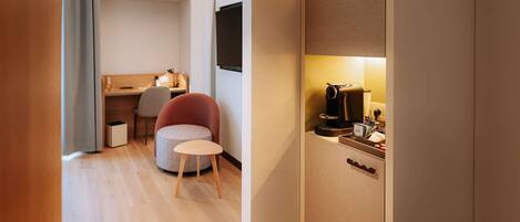 Una habitación de hotel moderna con suelo de madera, un escritorio con una silla, una pequeña mesa redonda, un armario y una cafetera.