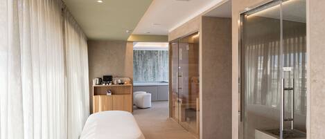 A modern hotel room with a large bed, a seating area, a glass-enclosed shower, and a view of the city.