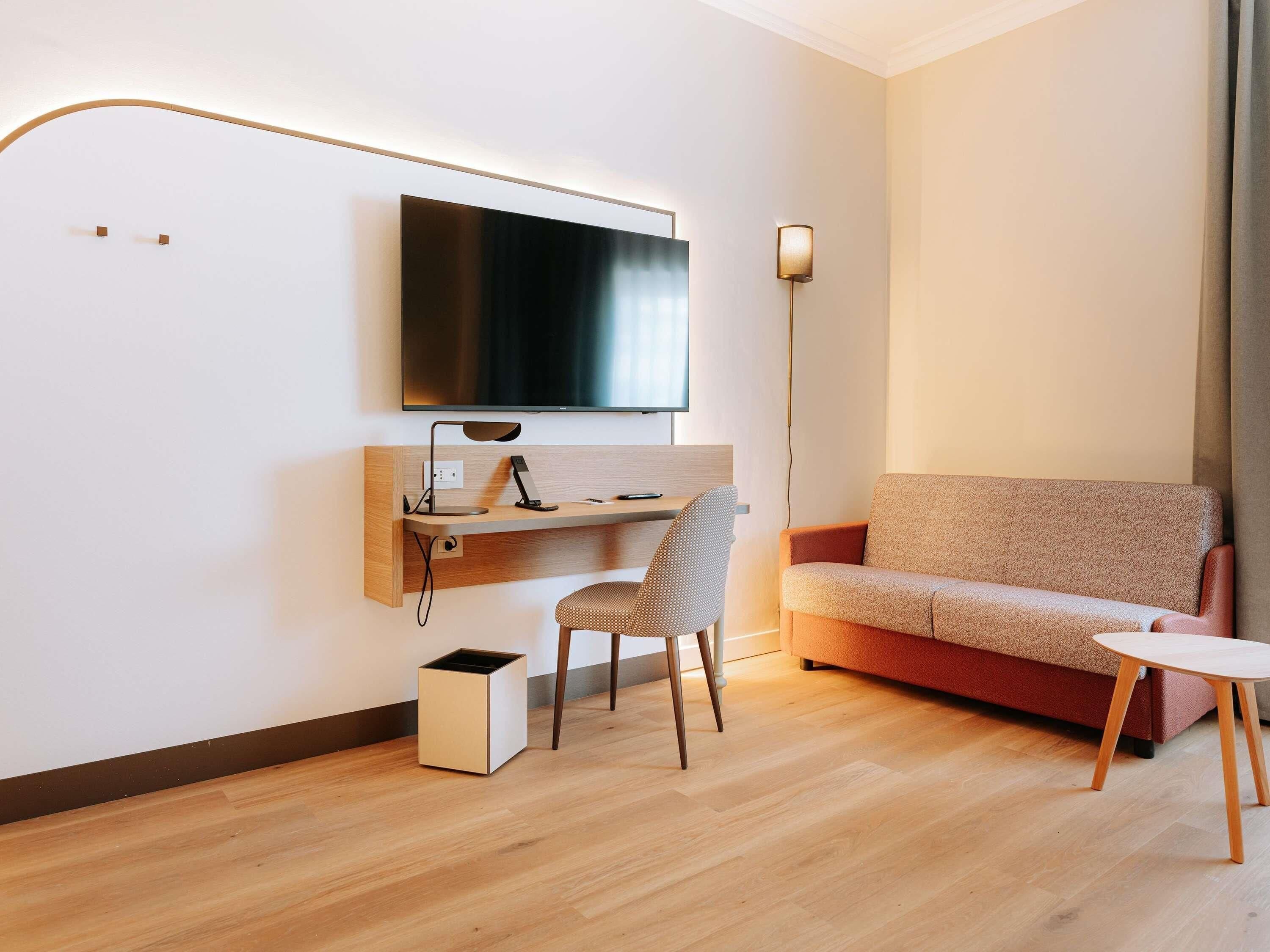 A modern hotel room with a flat-screen TV mounted on the wall, a desk with a lamp, a patterned chair, a sofa, and a small side table.