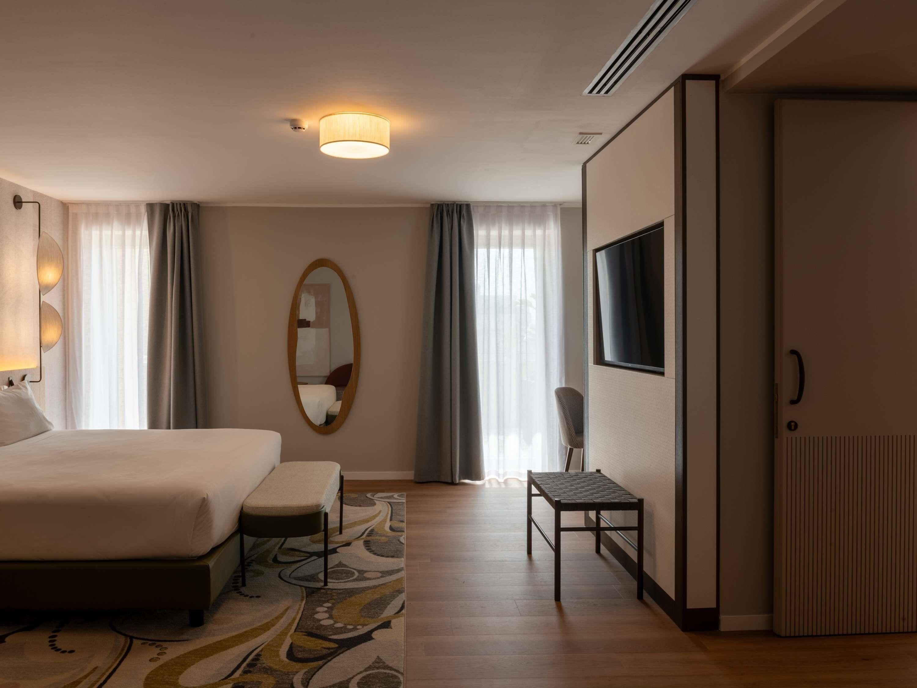 A hotel room with a bed, a chair, a TV, a mirror, and a window with curtains.