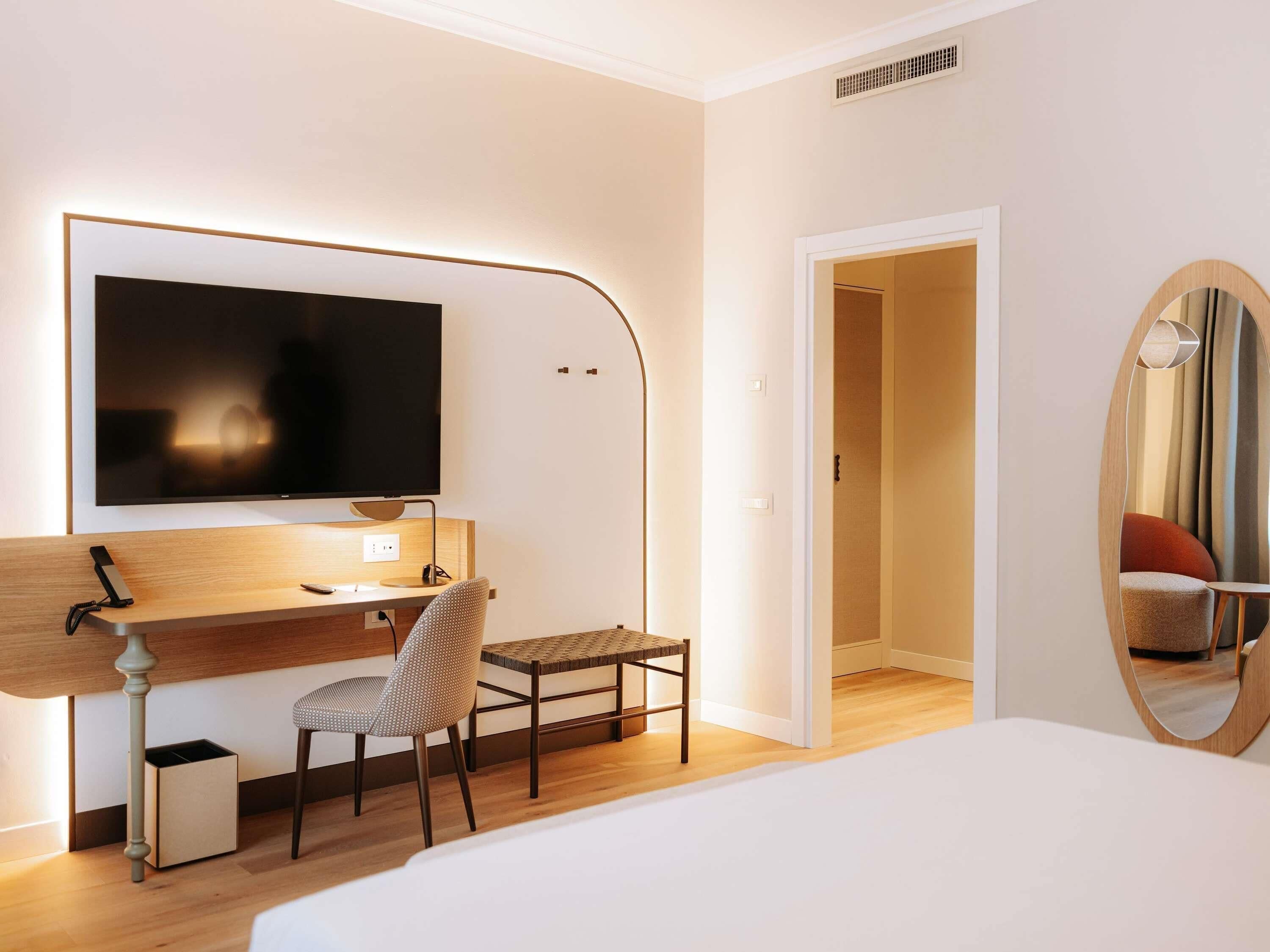 A modern hotel room with a bed, a desk with a chair, a TV, and a mirror.