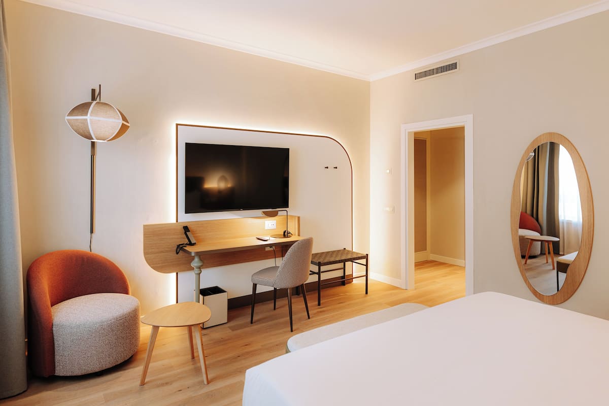 Ein modernes Hotelzimmer mit einem Bett, einem Schreibtisch mit Computer, einem Sessel, einer Lampe und einem großen Spiegel.
