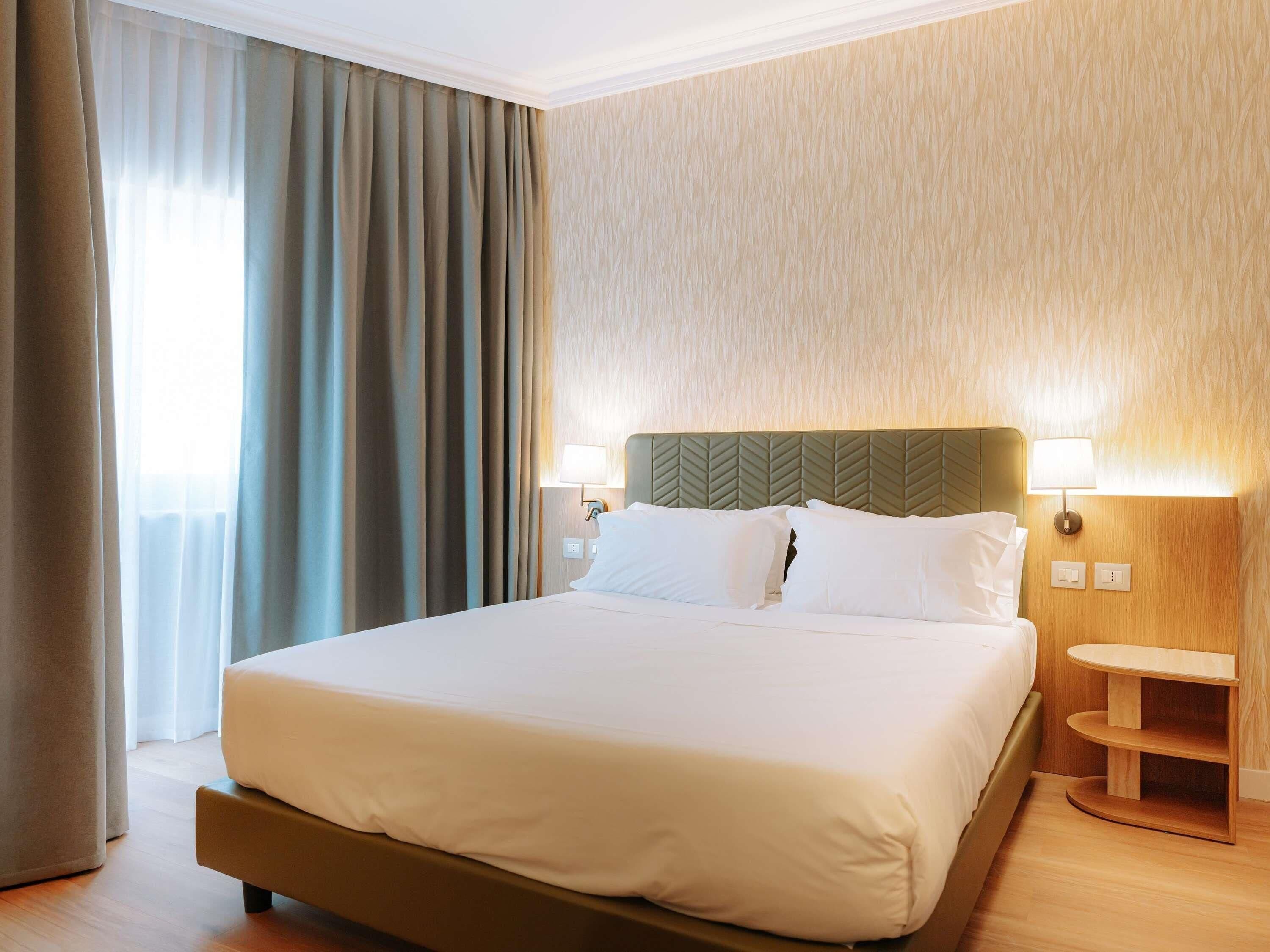 Una habitación de hotel con cama, cortinas, cabecero, lámparas fijadas a la pared y mesita de noche de madera.