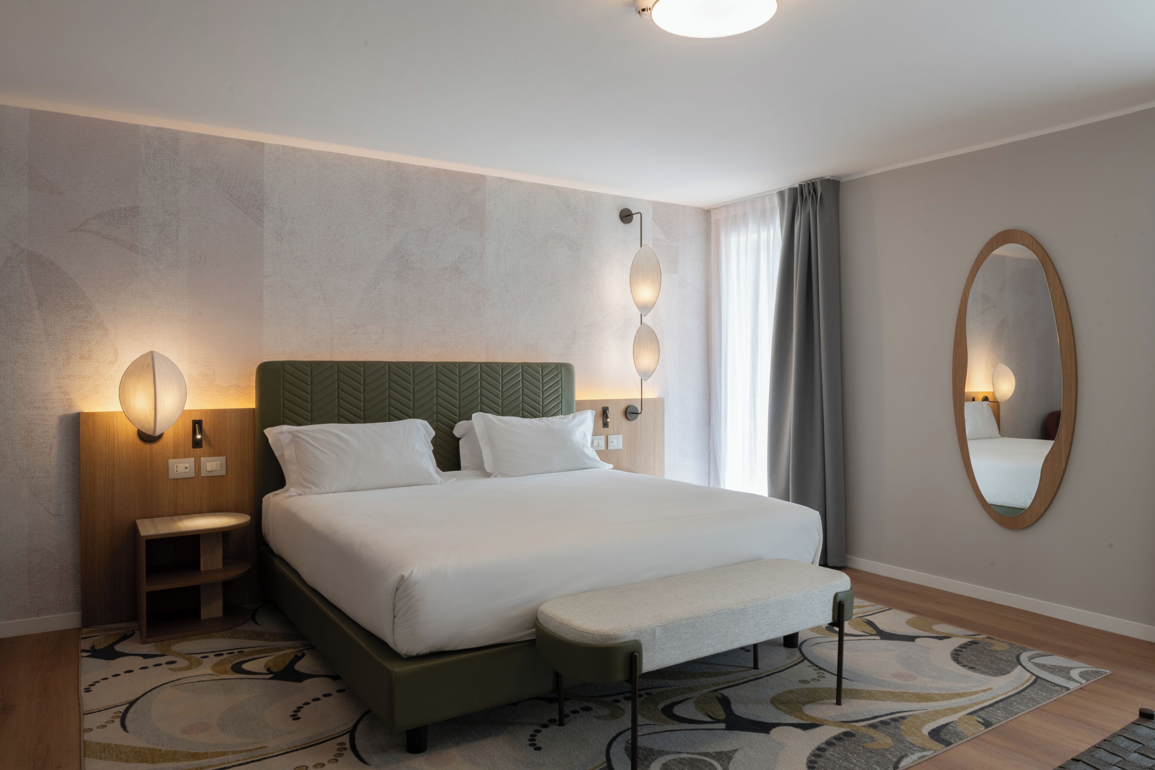 A hotel room with a bed, bedside tables, a bench, a wall-mounted mirror, and a patterned rug.