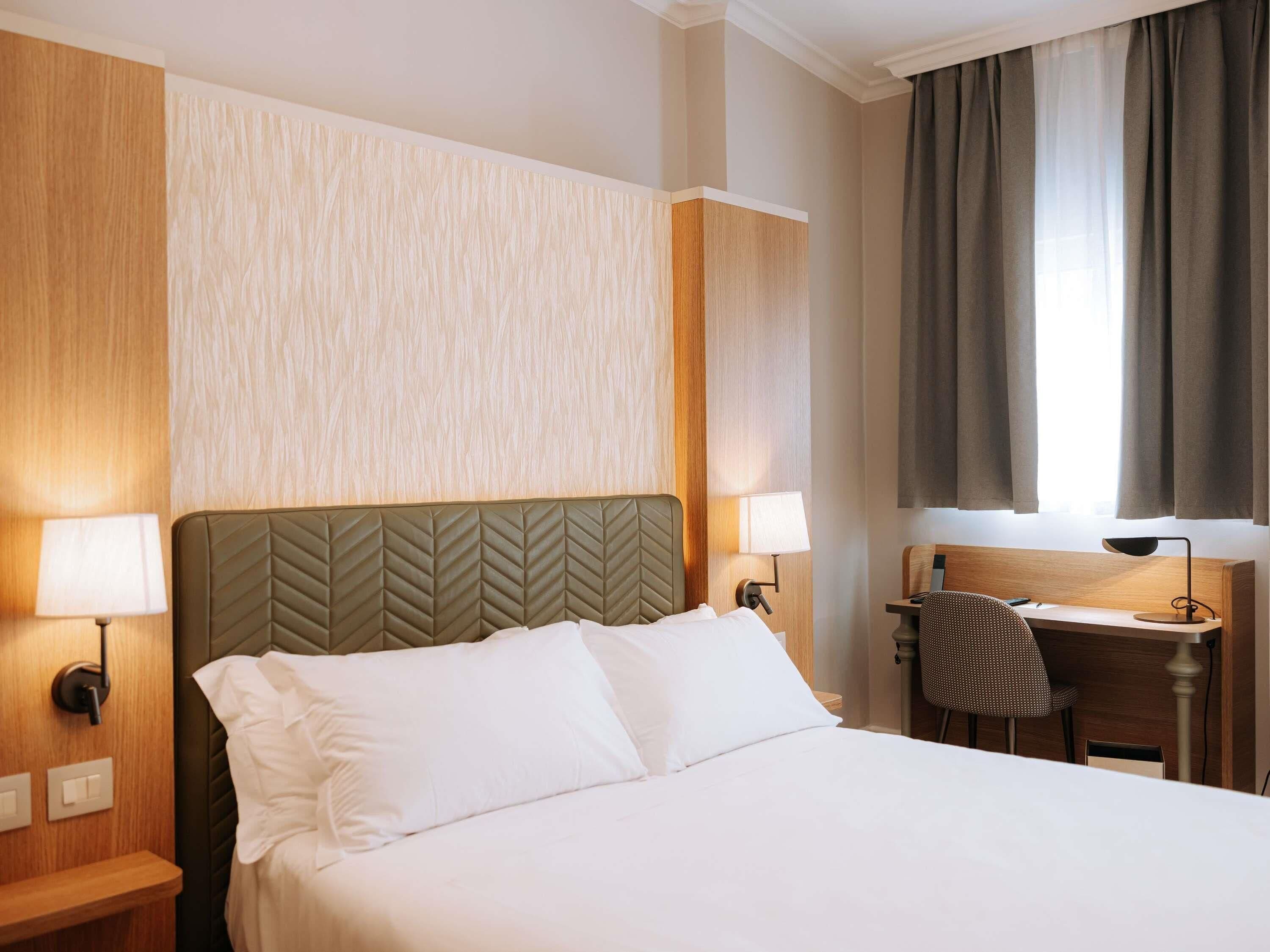 A hotel room with a bed, two bedside lamps, a desk with a chair, and a window with curtains.