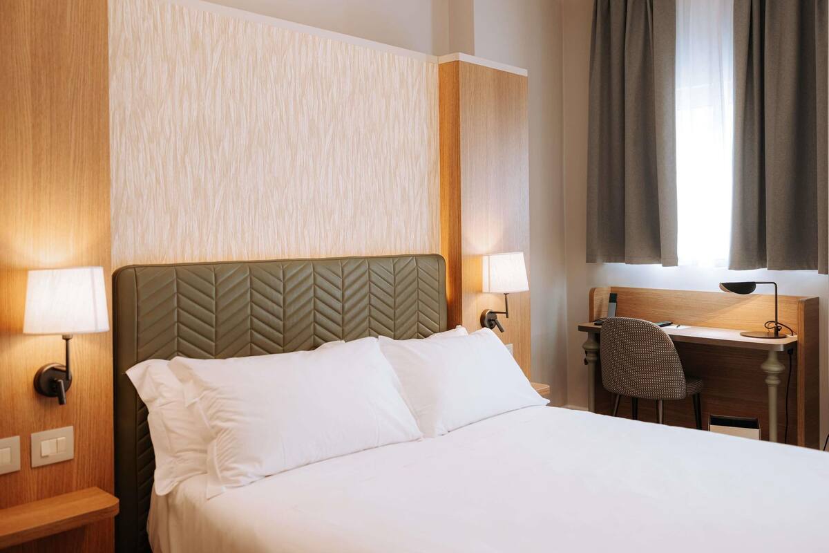 Ett hotellrum med en säng, två sänglampor, ett skrivbord med en stol och ett fönster med gardiner.