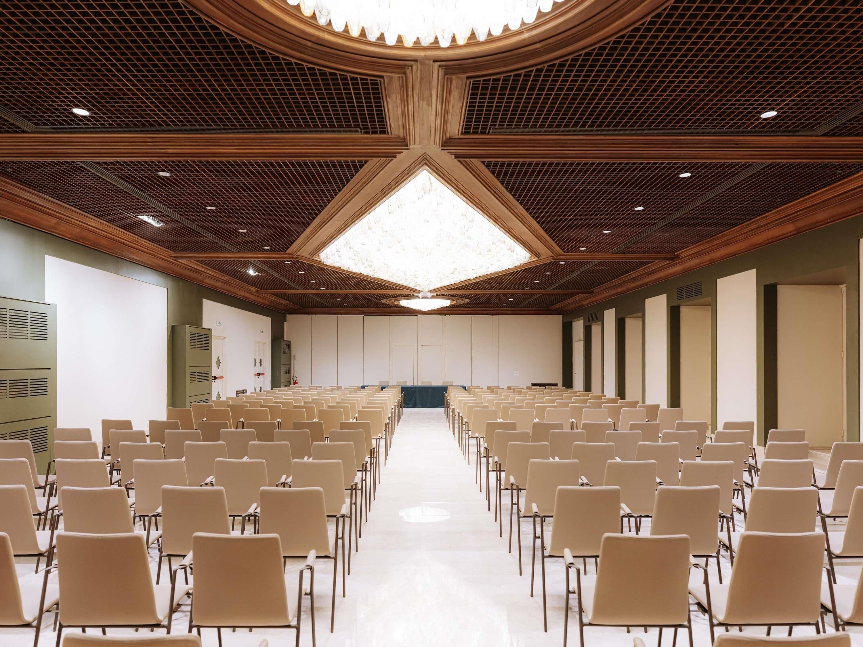 A conference room with rows of chairs, a high ceiling with a geometric pattern, and a large skylight.
