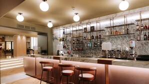 Uma área de bar com parede de mármore, diversas garrafas de bebidas e três banquetas.