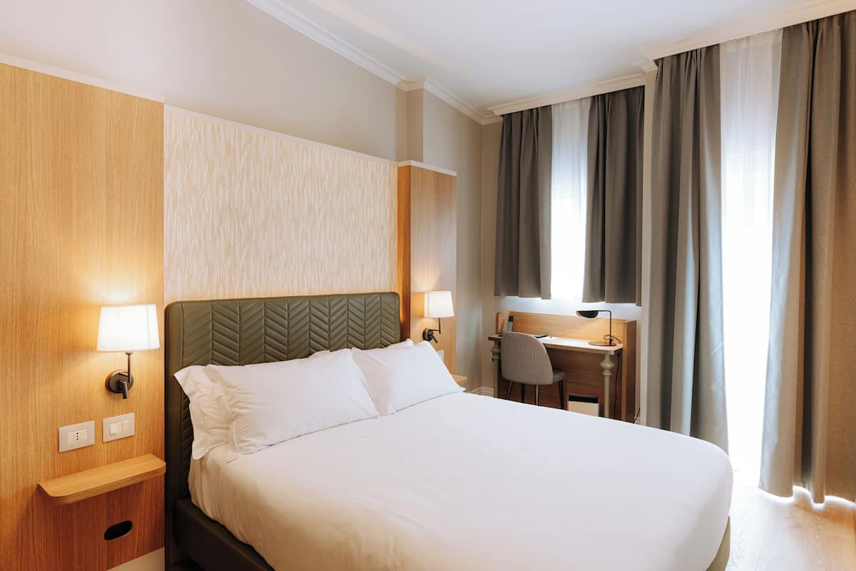 Ein Hotelzimmer mit einem großen Bett, einem Schreibtisch und einem Stuhl. Zwei Wandlampen und ein Fenster mit Vorhängen sind vorhanden.
